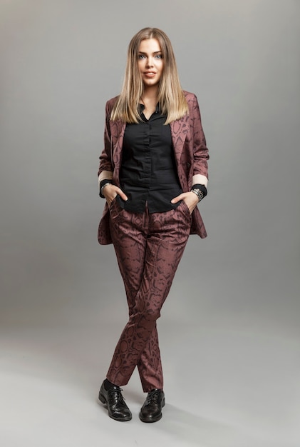 Beautiful young woman in a trouser suit. A spectacular blonde stands and holds her hands in her pockets. Business success. Gray background. Vertical.