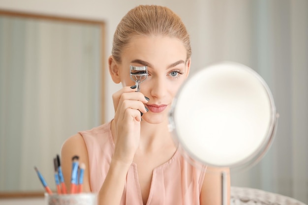 Beautiful young woman using eyelash curler at home