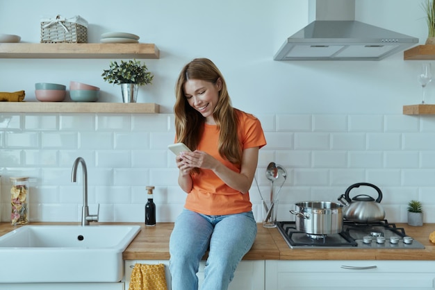 Beautiful young woman using smart phone while sitting at the kitchen counter