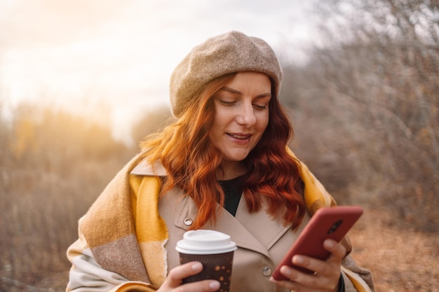 Beautiful young woman using smartphone and drinking coffee takeaway at city park