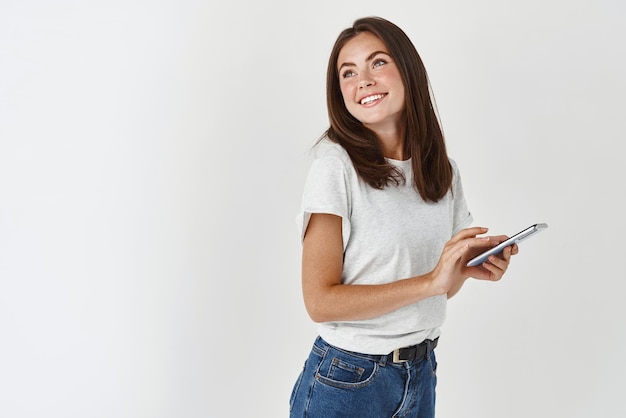 Beautiful young woman using smartphone smiling and dreamy looking at upper left corner logo standing over white background