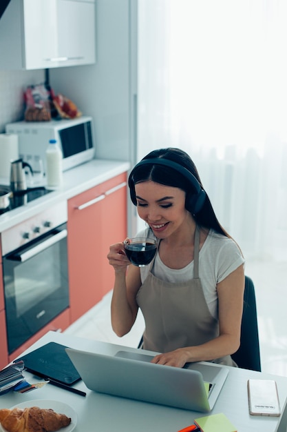 Beautiful young woman wearing an apron typing on the laptop keyboard and holding a glass cup