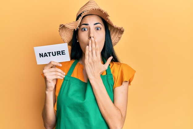 Beautiful young woman wearing gardener apron holding nature word covering mouth with hand, shocked and afraid for mistake. surprised expression