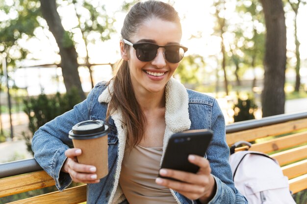 Beautiful young woman wearing jacket sitting on a bench, drinking takeaway coffee, using mobile phone