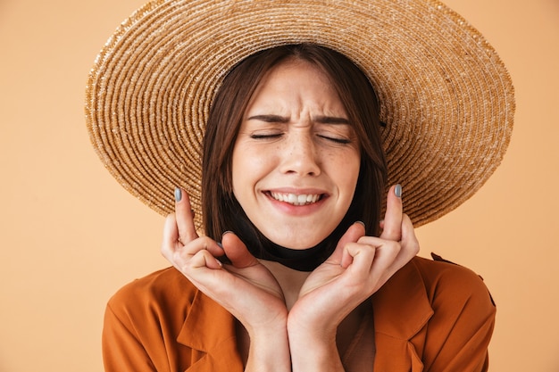 Beautiful young woman wearing straw hat standing isolated over beige wall, holding fingers crossed for good luck