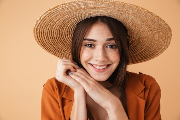 Beautiful young woman wearing straw hat standing isolated over beige wall, posing