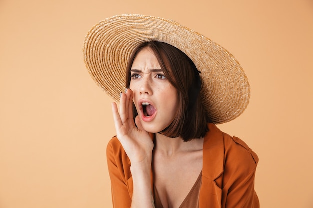 Beautiful young woman wearing straw hat standing isolated over beige wall, screaming