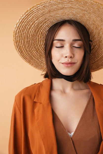 Beautiful young woman wearing straw hat and summer outfit standing isolated over beige wall, posing