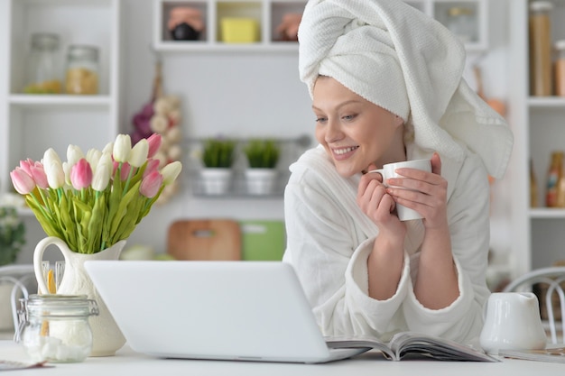 Beautiful young woman wearing a white bathrobe with laptop and drinking tea at home