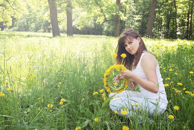 Beautiful young woman weaves a wreath of dandelions