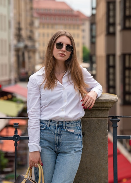 A beautiful young woman in a white blouse and blue jeans and sunglasses walks along a city street holding a handbag Girl with long dark ha