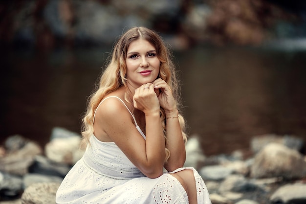 Beautiful young woman in a white dress in the mountains near the river on the background of a waterfall