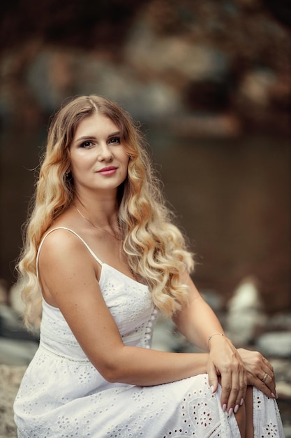 Beautiful young woman in a white dress in the mountains near the river on the background of a waterfall