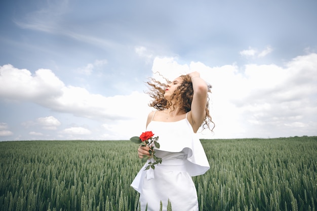 Beautiful young woman in the white dress standing in the field with rose in her hand