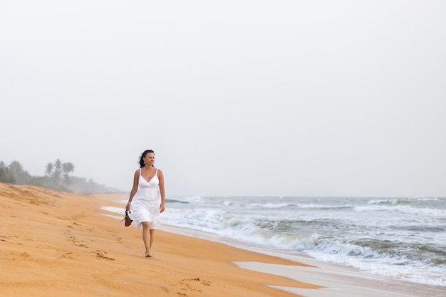 Beautiful young woman in white dress walking on the sandy beach