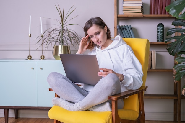 Beautiful young woman in white hoodie using laptop sitting on yellow armchair at home.