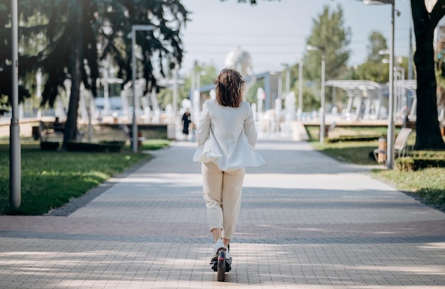 Beautiful young woman in white suit is riding to work on her electric scooter in city parkland