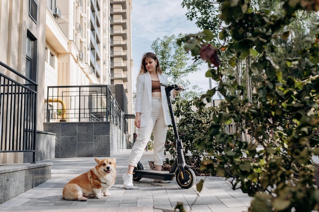 Beautiful young woman in white suit standing with her electric scooter and COrgi Dog near modern building in city and looking away