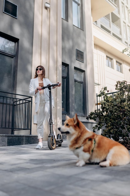 Beautiful young woman in white suit standing with her electric scooter and Corgi Dog near modern building in city and looking away