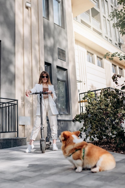 Beautiful young woman in white suit standing with her electric scooter and COrgi Dog near modern building in city and looking away