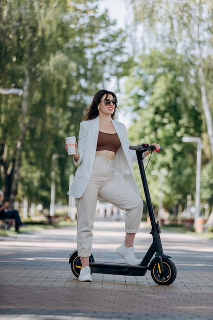 Beautiful young woman in white suit and sunglasses standing with her electric scooter in city parkland and drinking coffee