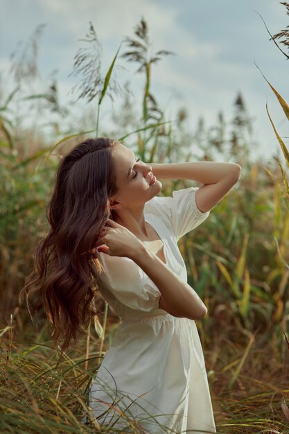 Beautiful young woman in white summer dress is sitting in tall grass in rural field Portrait of romantic girl at sunset warm sun natural beauty woman