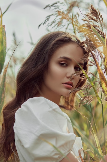 Beautiful young woman in white summer dress is sitting in tall grass in rural field Portrait of romantic girl at sunset warm sun natural beauty woman