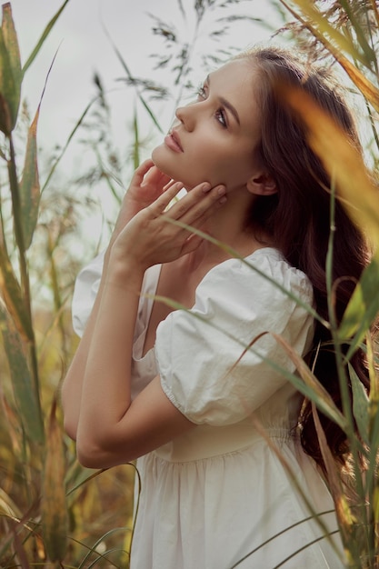 Beautiful young woman in white summer dress is sitting in tall grass in rural field Portrait of romantic girl at sunset warm sun natural beauty woman