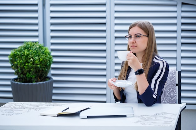 Beautiful young woman in a white T-shirt is drinking coffee and working on a laptop and smiling while sitting outdoors in a cafe. Young woman laptop for work. Female freelancer working on a laptop