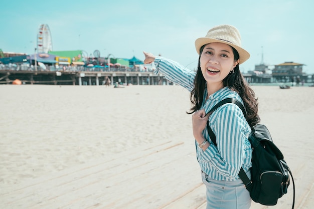 Beautiful young woman with backpack having fun at amusement park. relaxation in spring sandy beach concept. pretty girl traveler in straw hat looking face camera smiling point theme park on sunny day