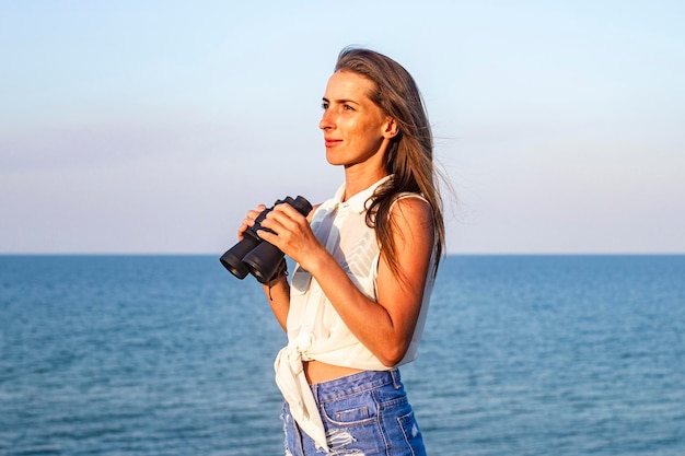 Beautiful young woman with binoculars on the cliff looks at the sea