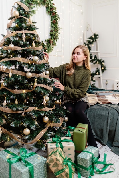 A beautiful young woman with blond hair sits near a Christmas tree and decorates with toys New Years is soon Christmas atmosphere in a cozy home