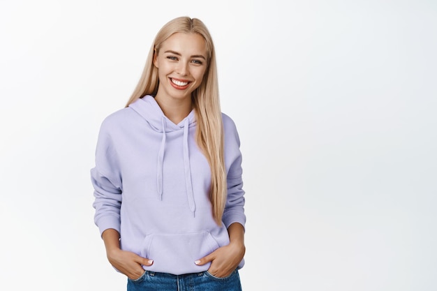 Beautiful young woman with blond hair white smile standing in casual hoodie holding hands in pockets posing over white background