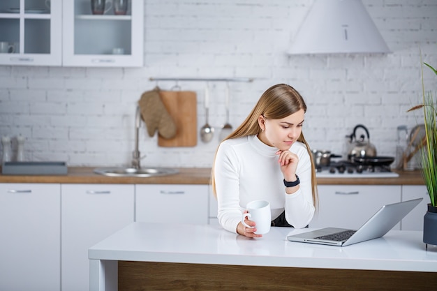 A beautiful young woman with blond hair in a white turtleneck sweater stands in a white kitchen and works at a laptop, she drinks coffee. Work remotely from home