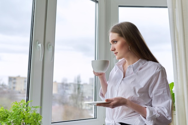 Beautiful young woman with blond long hair holding cup coffee near window