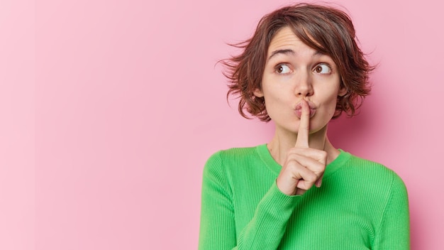 Photo beautiful young woman with bob hairstyle makes hush gesture shows silence sign wears green jumper focused mysterious away isolated over pink background blank copy space for your advertisement