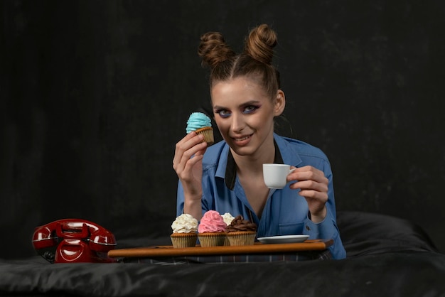 Photo beautiful young woman with bright makeup and bun hairstyle drinks coffee with muffins decorated with multicolored cream