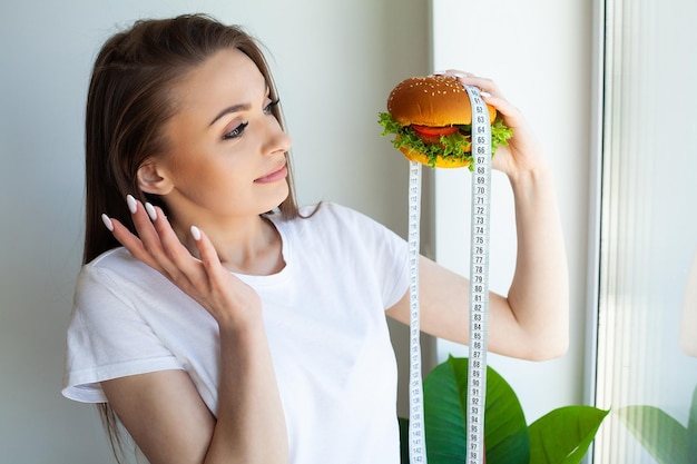 Beautiful young woman with burger and measuring tape while dieting