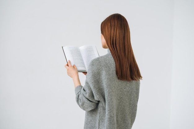 Beautiful young woman with dark long hair in grey knitted sweater reading book on the white background