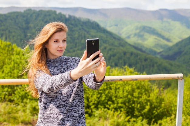 Beautiful young woman with long hair is doing selfie on her phone on the background of green nature