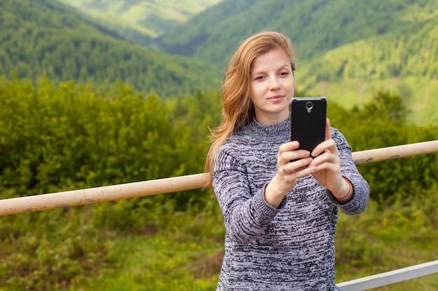 Beautiful young woman with long hair is doing selfie on her phone on the background of green nature