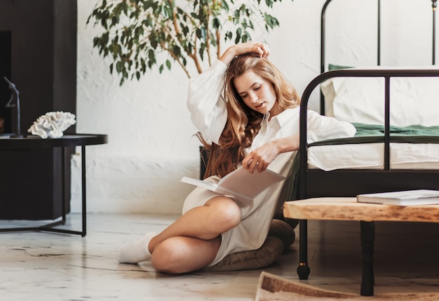 Beautiful young woman with long hair reading a book sitting in bedroom with white sheets