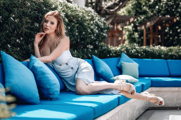 Beautiful young woman with long legs is lying on the blue sofa near the leaves