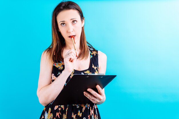 Beautiful young woman with pen and notepad pensive or doubtful on blue background