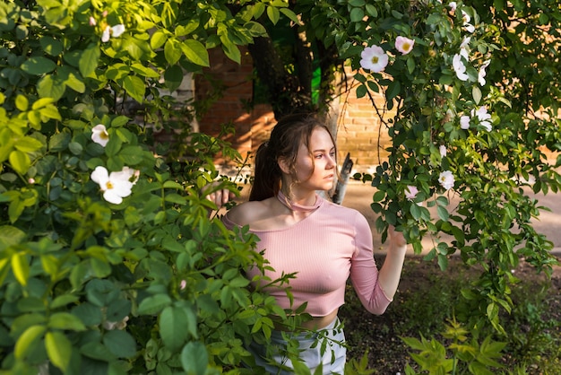 Beautiful young woman with perfect skin in a pink outfit posing near blooming roses in the garden