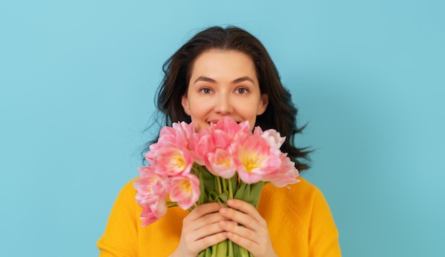 Beautiful young woman with pink flowers tulips in hands on blue wall background