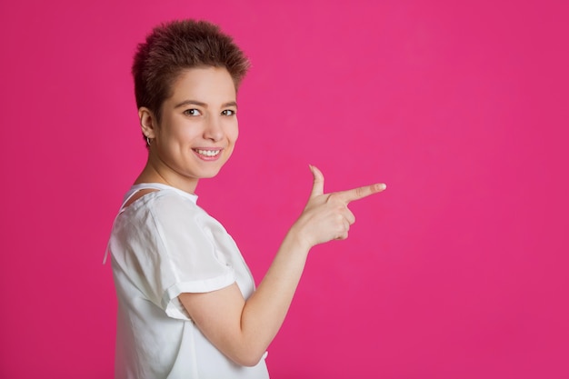 beautiful young woman with short hairdo in a white t-shirt on a pink wall pointing a finger to the side