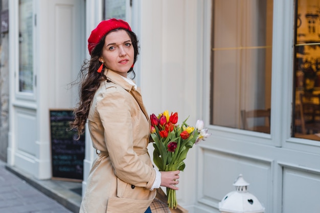 Beautiful young woman with spring tulips flowers bouquet at city street. Happy girl walking outdoors. Spring portrait of pretty female in old town