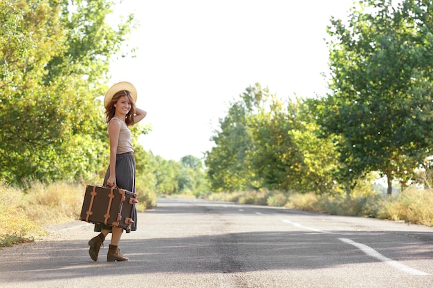 Beautiful young woman with suitcase standing on road