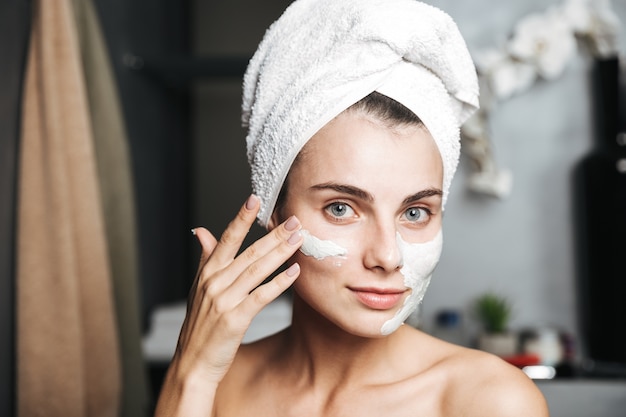 Beautiful young woman with towel wrapped around her head applying face mask at the bathroom